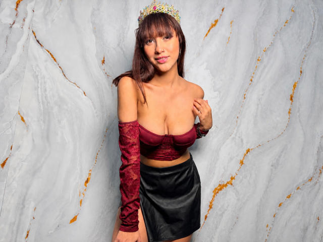 HanaJoud, con un seno di media grandezza, indossa una maglia rossa di velluto con una spalla scoperta, una gonna nera e una tiara dorata. Ha lunghi capelli castani e un sorriso seducente. Il suo braccio è appoggiato sul petto..