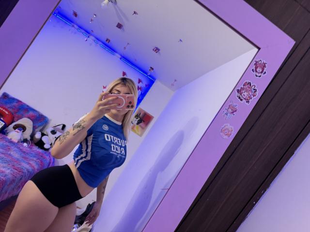 SammyViolet in a blue crop top and black shorts, showing off her tats and ass in a mirror selfie, holding a pink phone. She's got blonde hair and a cute face.