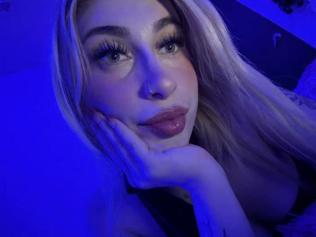 SammyViolet with blue-lit face, pouty lips, long blonde hair, thick eyelashes, nose ring, and hand resting on cheek, wearing black top, looking at camera.