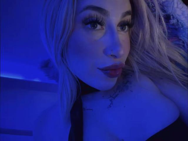 SammyViolet with blonde hair, blue-lit face, nose ring, long eyelashes, red lipstick, tattoo on chest, black strap dress, slight smile, looking at camera, blue-toned selfie.