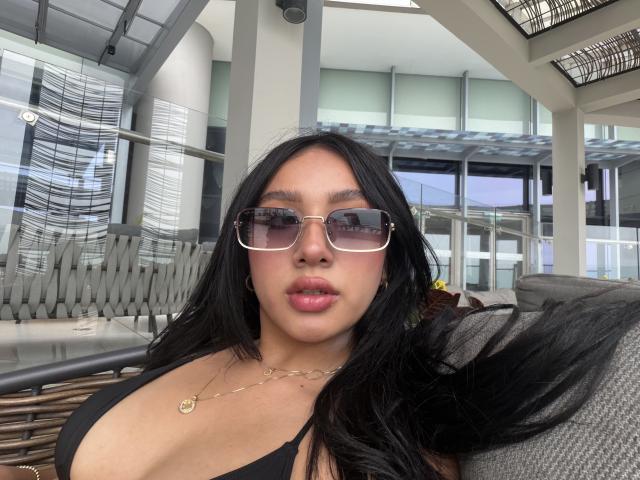 LillyTaylor with long black hair, wearing sunglasses, a black top showing her big tits, and a gold necklace, sitting outdoors on a gray couch.