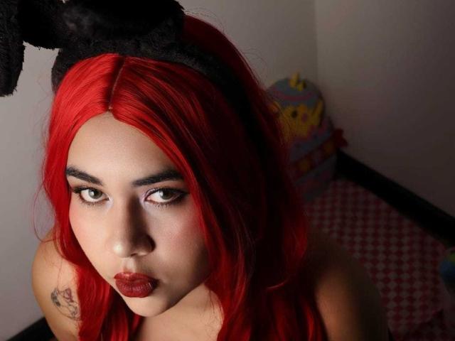 LilithVelvett with red hair, black fuzzy bunny ears, heavy eyeliner, and red lipstick, looking up with pouty lips, tattoo on left shoulder, in a dimly lit room.