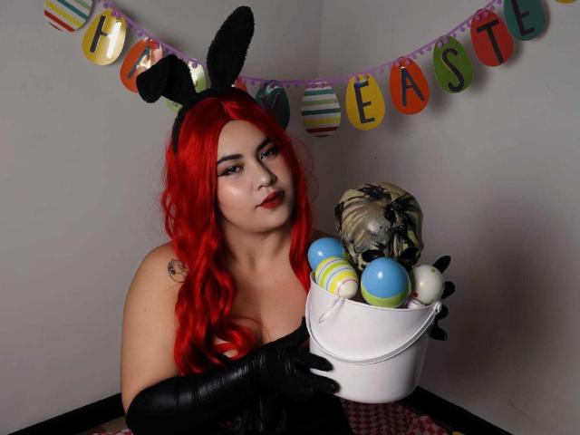 LilithVelvett with red hair, black bunny ears, black gloves, holding a basket of colorful Easter eggs and a stuffed bee. Her tits are visible, she's looking at the camera.