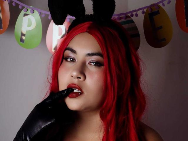 LilithVelvett with red hair, black bunny ears, and glossy black glove, biting her red lipstick with her finger, looking seductive.
