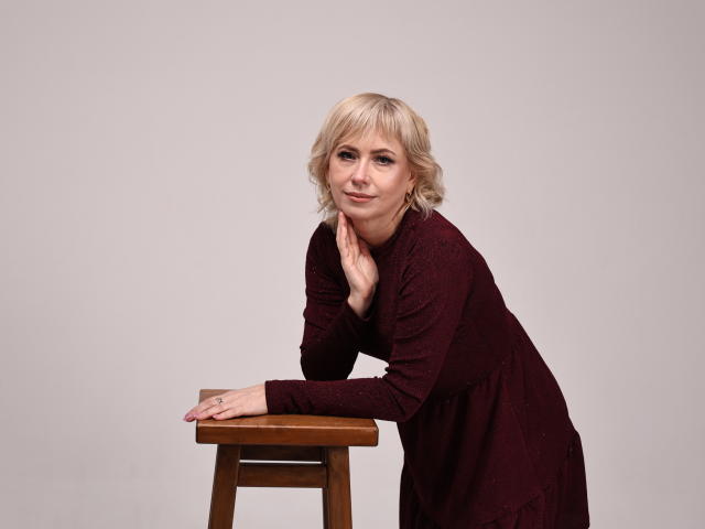 Lauren Wishy mit kurzen, blonden Haaren, in einem burgunderfarbenen Kleid, lehnt an einem hölzernen Hocker, Hand am Kinn, lächelnd, rote Fingernägel, helle Haut, ungezwungene Studioaufnahme..