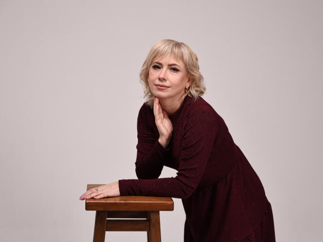 LaurenWishy, blonde, wearing a maroon dress, leans on a wooden stool with one hand under her chin, looking slightly up, smiling subtly. Her ass is not visible.
