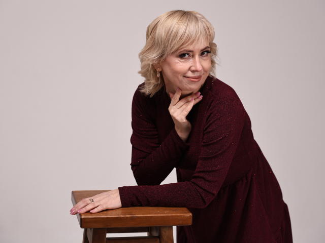 LaurenWishy, blonde, vêtue d'une robe rouge scintillant, est appuyée sur un tabouret en bois, sa main posée sur son menton, et elle sourit doucement. Ses seins sont petits, et elle est mignonne..