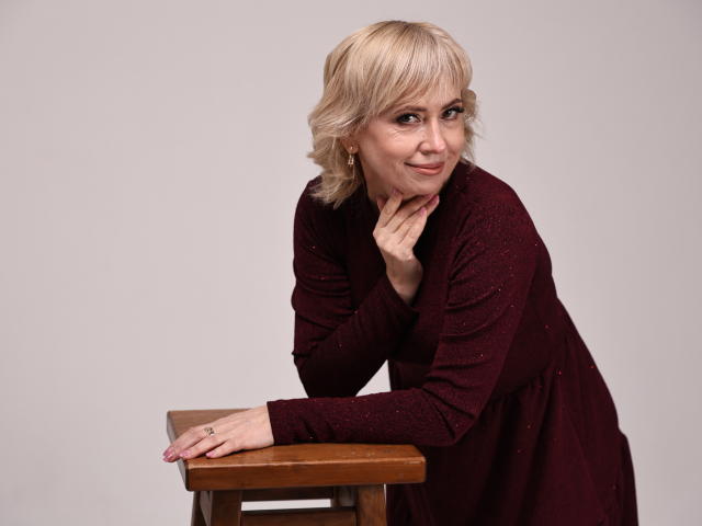 LaurenWishy with blonde hair, wearing a maroon dress, leaning on a wooden stool, smiling, hand near chin, ring visible, pink nails, soft light, casual pose.