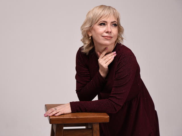 LaurenWishy with blonde hair, wearing a sparkly maroon dress, leans on a wooden stool, hand on chin, smiling softly. Boobs visible under dress, pink nails, subtle makeup.