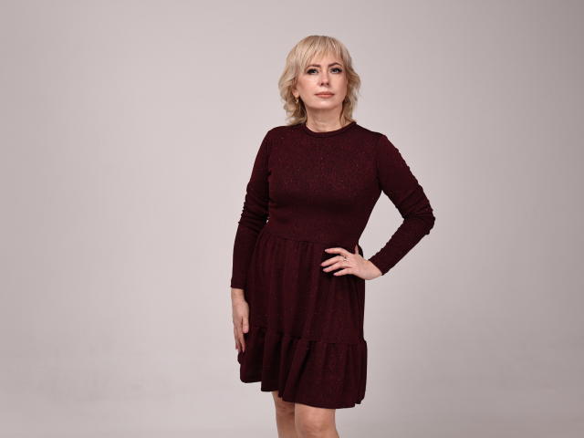 LaurenWishy stands with one hand on her hip, wearing a dark red, long-sleeved dress with a lace pattern and ruffled skirt, looking slightly to the side.