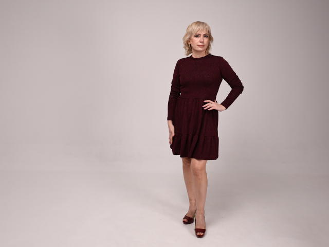 Lauren Wishy, avec ses cheveux blonds, portant une robe rouge foncé, debout avec une main sur sa hanche, et des talons hauts marron, regardant légèrement vers la droite..