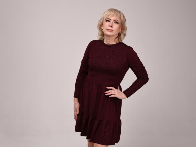 LaurenWishy with blonde hair, wearing a dark red, long-sleeve dress with a ruffled bottom, standing with one hand on her hip, looking slightly to the side.