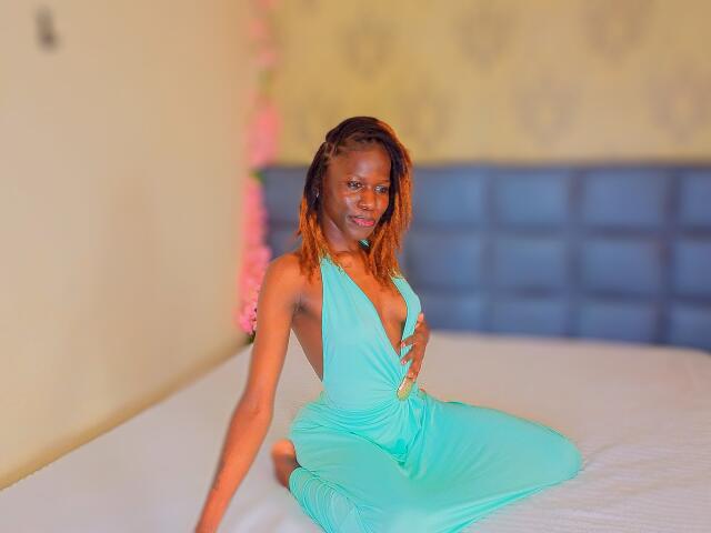 KharlaQueen in a turquoise dress with a deep V, sitting on a bed, showing her small tits, smiling, with orange-brown braided hair, on a white bed.