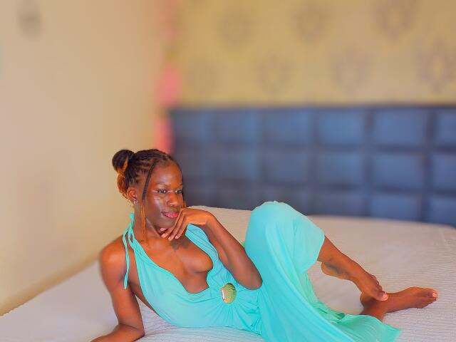 KharlaQueen with dark skin, small tits, and a sexy smile, wearing a teal, halter dress, lying on a bed, one hand on her chin, the other on her thigh.