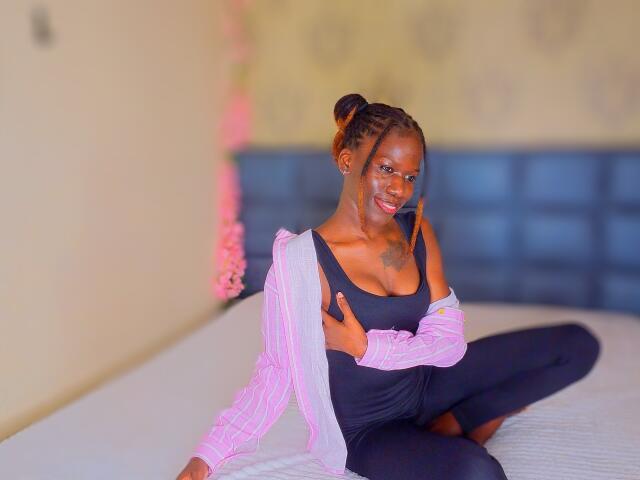 KharlaQueen with dark skin, small boobs, and a smile, sitting on a bed in a black top and pink cardigan, hands on her chest, legs crossed, looking at camera.