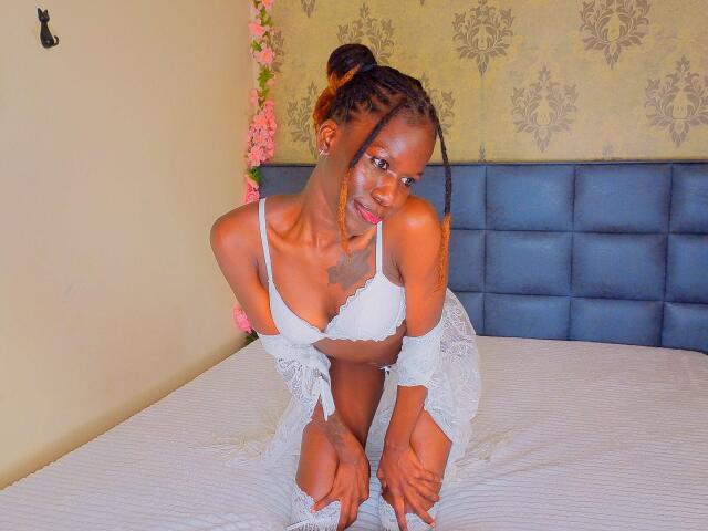 KharlaQueen is kneeling on a bed wearing white lace lingerie, her small tits and dark skin visible, with braided hair and a cute smile, hands on the bed.