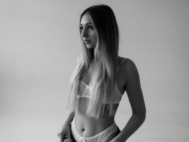 Celinda Feig, con cabello rubio largo, vestida con un sujetador blanco transparente y bombachas de encaje, de pie y mirando hacia un lado, mostrando senos pequeños y un cuerpo delgado y tonificado. Foto en blanco y negro..