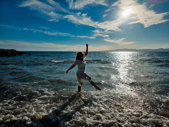 SallyVerda, with her ass and back to the camera, dances in the shallow water with one arm raised, wearing a white dress, under a bright sunlit blue sky.