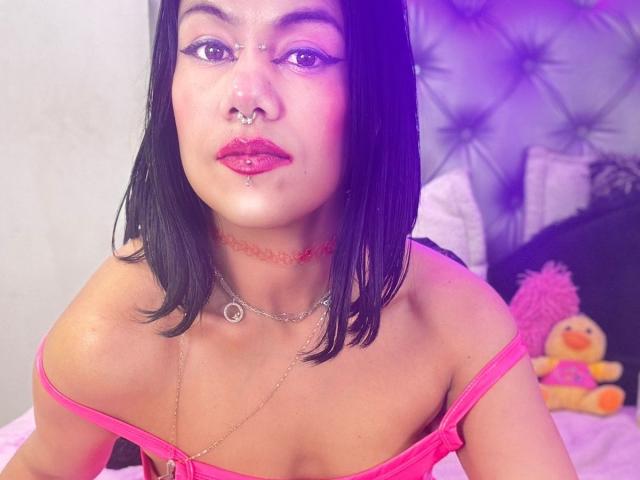 TarahDustin with black bob, pink lipstick, septum piercing, and lip piercing, wearing pink off-shoulder top, chains, and red choker, sitting on pink bed with stuffed duck.