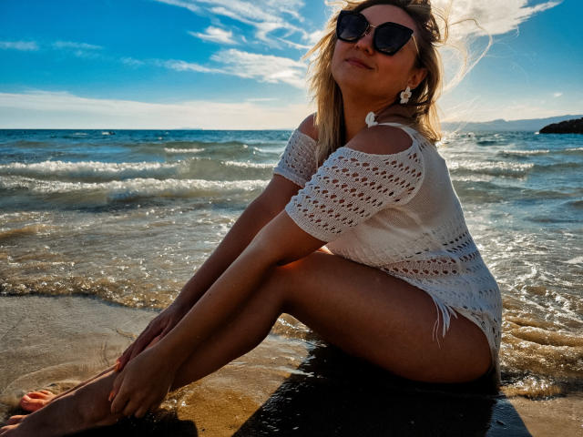 SallyVerda with blonde hair, wearing sunglasses, a white crocheted off-shoulder top, and white fringed bottoms, sitting on the wet beach, legs crossed, sunlight on her ass.