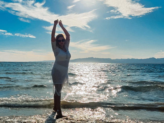 SallyVerda stands in the water with arms raised, wearing a white, see-through dress, sun shining, ocean waves around her legs, blue sky, and distant mountains in the background.