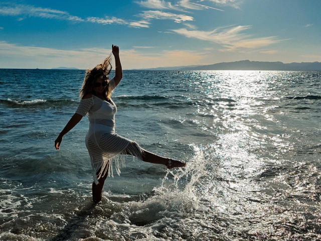 SallyVerda with long hair, white fishnet dress, standing in ocean, raising right arm, left leg in water, sunlight reflecting, waves splashing, clear sky, distant mountains.