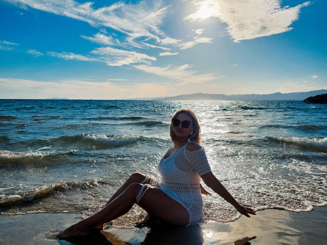 SallyVerda mit Brüsten, Hintern und Vulva, sitzend im feuchten Sand, trägt ein weißes, gehäkeltes Kleid, die Sonne scheint, blaues Meer, kleine Wellen, klarer Himmel, dunkle Sonnenbrille, blondes Haar, entspannt..