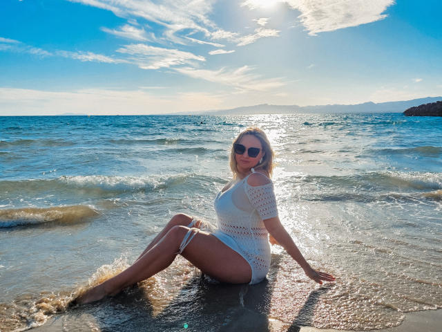 SallyVerda with sun-kissed skin, wearing a white, see-through knit dress, sitting in shallow water, her ass and tits visible, blonde hair, sunglasses, and a sexy smile.