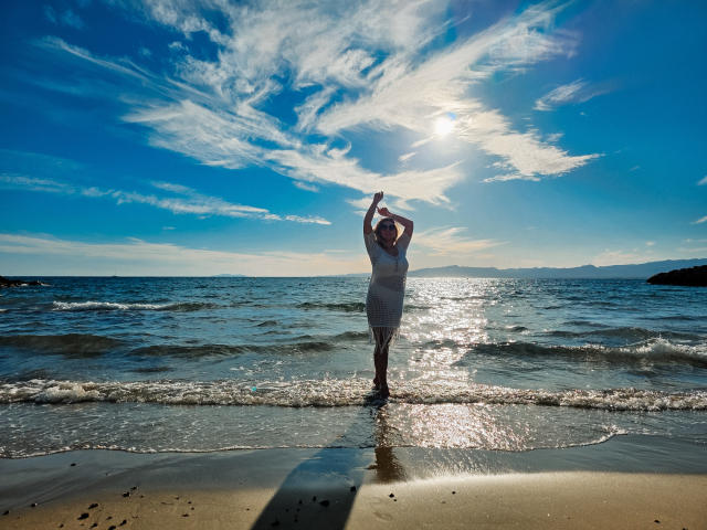 SallyVerda stands on a beach with arms raised, wearing a white dress, sunlight reflecting on her, ocean waves gently touching her feet, clear blue sky with wispy clouds.
