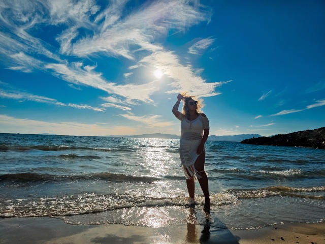 SallyVerda in einem weißen Kleid, stehend im Wasser, ihren Hut haltend, Sonnenlicht, das auf ihr reflektiert, blauer Himmel mit flauschigen Wolken, kleine Wellen, Strand, Felsen im Hintergrund..