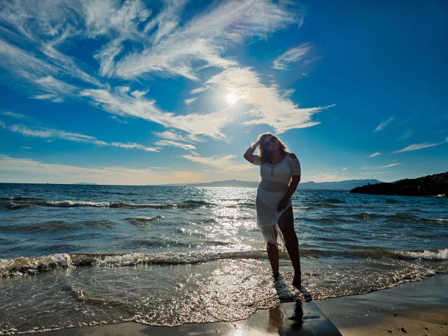 SallyVerda steht im flachen Wasser, trägt ein durchsichtiges weißes Kleid, die Sonne scheint auf sie, sie hat eine Hand im Haar, blauer Himmel mit weißen Wolken, die Sonne spiegelt sich in ihrem nassen Kleid..