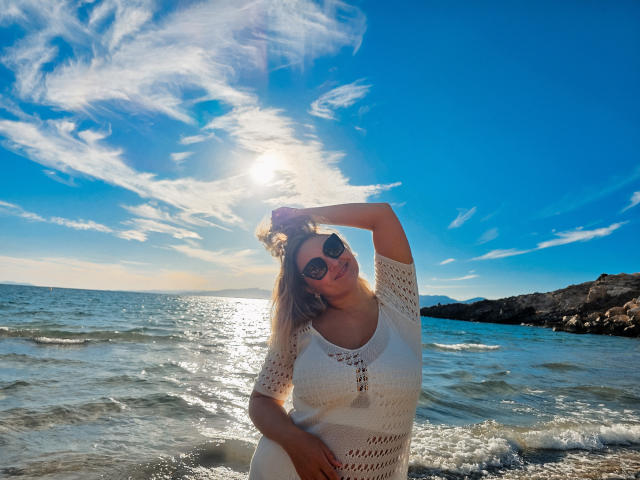 SallyVerda mit sonnengebleichtem, blondem Haar, trägt ein weißes, gehäkeltes Kleid, eine Sonnenbrille und hat eine Hand im Haar. Sie steht im flachen Meerwasser, der Himmel ist blau mit zarten Wolken, die Sonne steht hinter ihr..