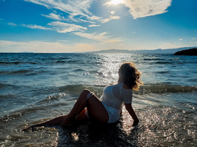 SallyVerda sitting in shallow water, back to camera, wearing a white, see-through, knitted top, sun reflecting off her ass, ocean, clear sky, sunlight, small waves.