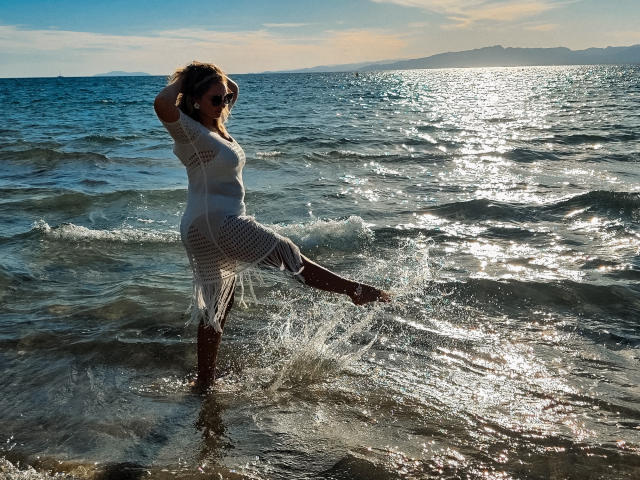 SallyVerda, standing in the ocean, wearing a white, see-through, mesh dress, with her leg raised, splashing water, sun shining, and her hands in her brown hair.