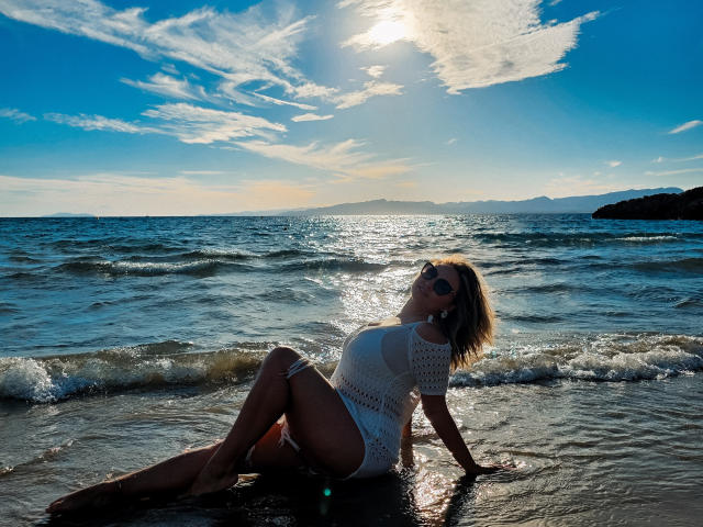 SallyVerda with her tits and ass in a white crochet dress, sitting in the wet sand at the beach, wearing sunglasses, sun shining, ocean and sky in the background.