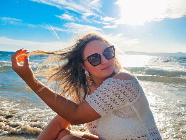 SallyVerda with tits covered by a white, off-shoulder, crochet top, holding her blonde hair, wearing black sunglasses, and white earrings, sitting on a pebbled beach, sun shining.