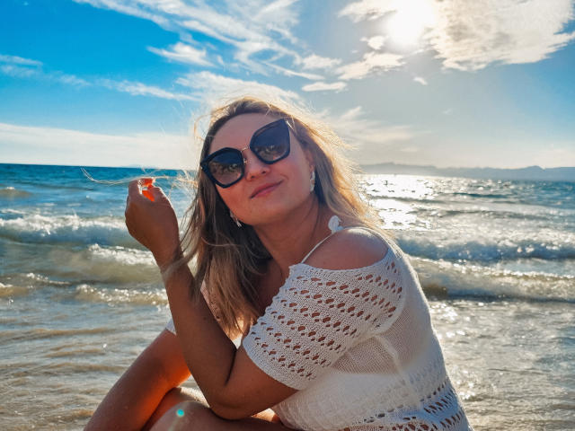 SallyVerda with long blonde hair, wearing dark sunglasses, white crochet top, sitting on beach, sun reflecting off water, holding hair, blue sky with clouds, smiling.