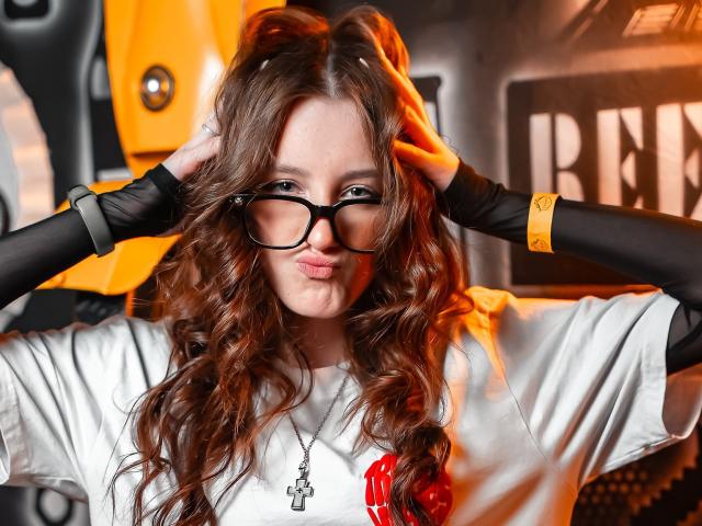 Leticia Ettl mit welligem, braunem Haar, schwarzen Brillen, voller Lippen, einem weißen Hemd, schwarzen Ärmeln, einem gelben Armband, den Händen im Haar, einer Halskette, vor einem orangefarbenen Hintergrund, die in die Kamera schaut, sexy..