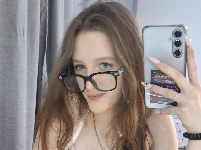 Leticia Ettl mit langen, braunen Haaren, die eine schwarze Brille trägt, ein Telefon vor ihrem Gesicht hält, lächelt, kleine, weiße Brüste zeigt und ein weißes Oberteil trägt..