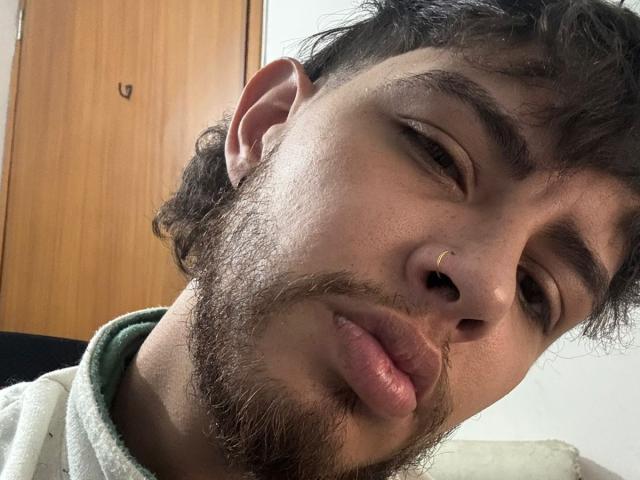 Sevven has curly dark hair, a beard, a nose ring, and puckered lips in a selfie. His eyes are half-closed, and he's wearing a green hoodie.