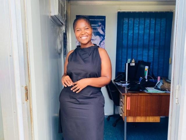 BettinaCori with dark skin, smiling, wearing a black sleeveless dress, standing in a doorway, hand on her slightly rounded belly, blue curtains in the background, office setting.