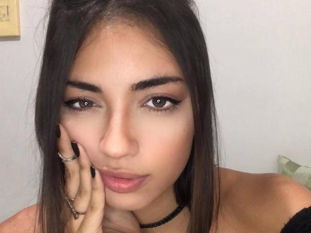 CameliaDeihl has brown eyes, long black hair, and a slightly pouty face with pink lips. She's touching her cheek with a ringed hand, wearing a black choker.