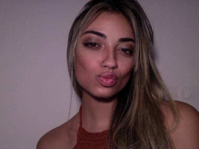 CameliaDeihl with pouty lips, nose ring, and long blonde hair, wearing a red halter top, looking slightly to the side with a subtle smile.