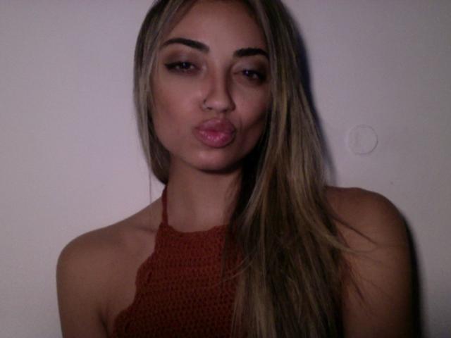 CameliaDeihl with pouty pink lips, nose piercing, long blonde hair, red crocheted halter top, and dark makeup, looking directly at camera.