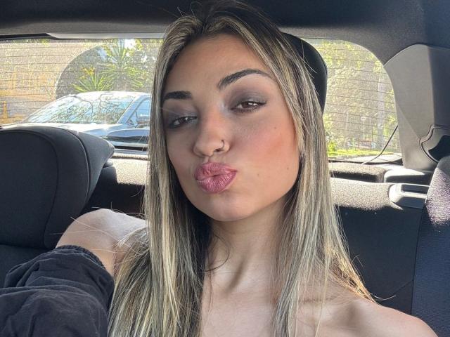 CameliaDeihl with pouty pink lips, nose ring, and long blonde hair, sitting in a car, wearing a black top, looking slightly to the side with a sultry expression.
