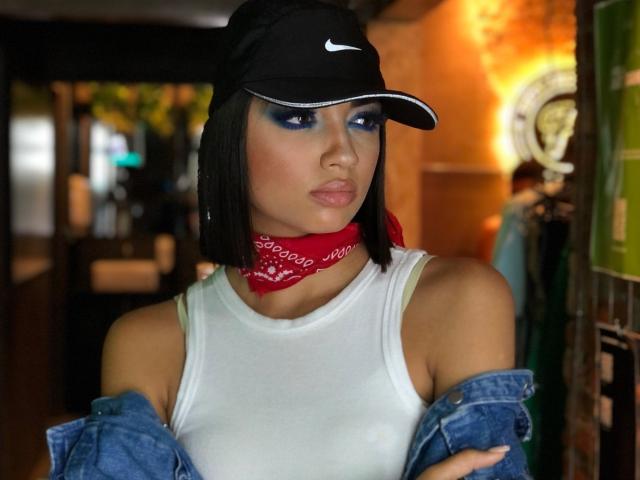 CameliaDeihl with blue eye makeup, black Nike cap, red bandana, white tank top, and blue denim off-shoulder, looking serious. She's got a short black bob and crossed arms.