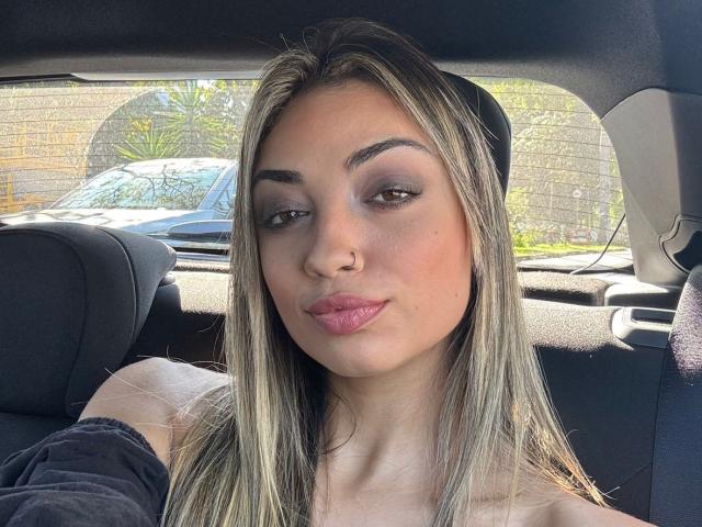 CameliaDeihl has blonde hair, dark eyebrows, blue eyeshadow, pink lips, a nose ring, and a slight smirk. She's in a car with black seats.