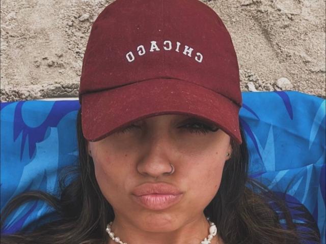CameliaDeihl with a maroon baseball cap, nose ring, and pearl necklace, pouting lips, brown eyes, and dark hair, lying on a blue patterned surface.