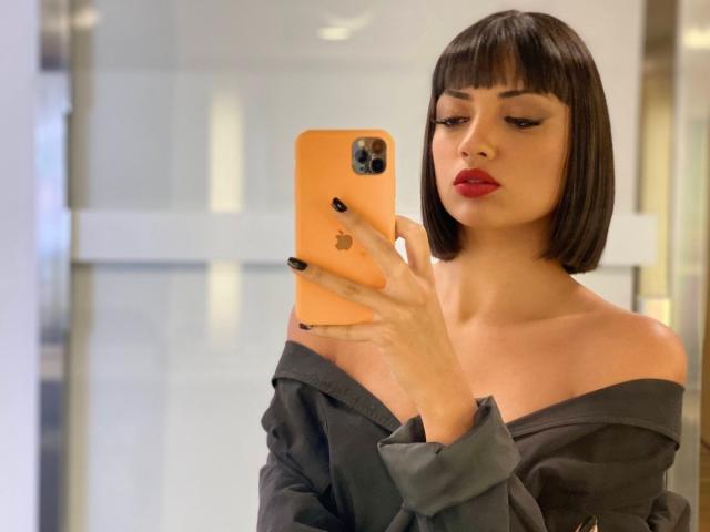 CameliaDeihl with a bob, red lipstick, black off-shoulder shirt, and black nails, holding an orange iPhone, taking a selfie. Her tits are covered by the shirt.