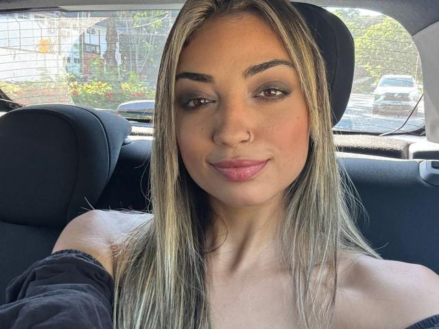 CameliaDeihl has blonde hair, brown eyes, a nose ring, and pink lips, sitting in a car with a black headrest, wearing a black off-shoulder top, smiling.